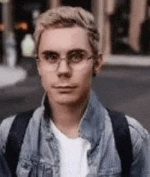 a young man wearing glasses and a denim jacket is standing on the street .