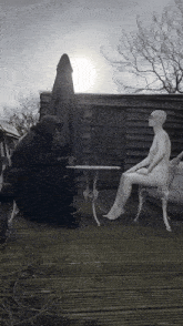 a man in a hood sits at a table with a naked mannequin
