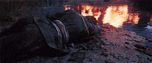 a man is laying on the ground in front of a river with a fire in the background