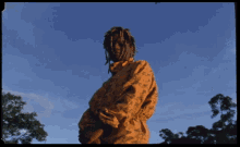 a man with dreadlocks stands in front of a blue sky with trees in the foreground