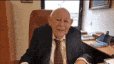 an elderly man in a suit and tie is sitting at a desk in front of a window .