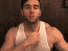 a man in a white tank top is standing in front of a wooden wall and looking at the camera .