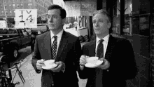 two men in suits and ties are walking down a sidewalk drinking coffee .