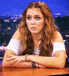 a woman is sitting at a table with her arms crossed and the words mysmiley on the bottom right
