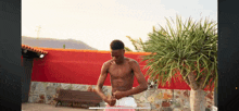 a shirtless man stands in front of a red wall and a palm tree