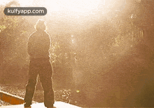 a man is standing on a bridge looking at the sun and the words kulfyapp.com are on the bottom right