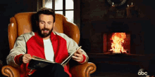a man in a red jacket is reading a book in front of a fireplace with abc written on the bottom right