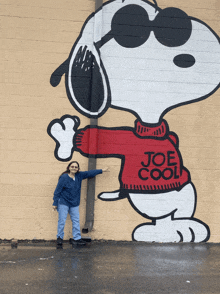 a woman stands in front of a mural of snoopy wearing a joe cool sweater