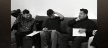 three men are sitting on a couch and one of them is wearing a black jacket with the word mcm on it