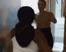 a woman is looking at herself in a mirror while a shirtless man is standing behind her .