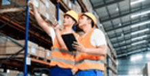 two workers are standing in a warehouse looking at boxes on shelves .