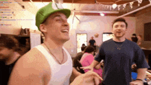 a man wearing a green hat and a white tank top laughs