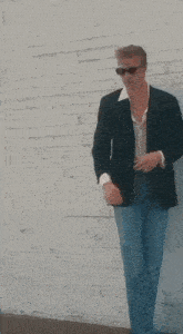 a man wearing sunglasses and a jacket is standing in front of a white brick wall .