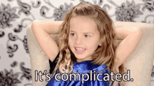 a little girl in a blue dress is sitting in a chair with her hands behind her head and says it 's complicated .