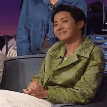 a man in a green jacket is sitting in a chair with his hands folded in front of him .