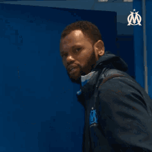 a man with a beard is covering his face with his hand while wearing a blue jacket with the letter m on it