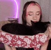 a woman with pink hair is holding a black cat in a floral bed