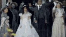 a bride and groom are standing in front of a crowd of people wearing old fashioned dresses .
