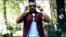 a man wearing sunglasses and a red vest is standing in a forest .