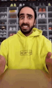 a man with a beard wearing a yellow hoodie is sitting at a table with his hands outstretched .