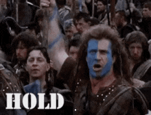 a man with blue paint on his face is standing in front of a crowd of people with their arms in the air .