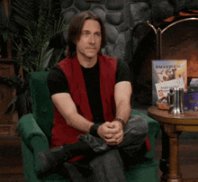 a man in a red vest sits in a chair with a daggerheart magazine behind him