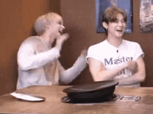two young men are sitting at a table laughing . one of the men is wearing a white shirt that says master .
