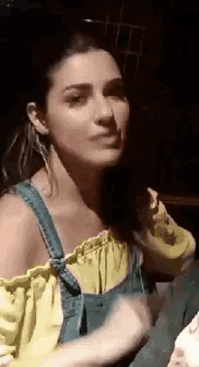 a woman in a yellow off the shoulder top and blue overalls is making a funny face