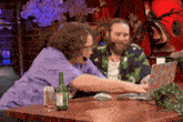 two men are sitting at a table looking at a laptop with a can of root beer on the table