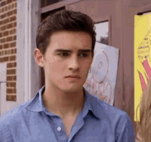 a young man in a blue shirt is standing in front of a brick building looking at the camera .