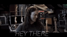 a man with long hair says " hey there " in front of a bunch of guitars