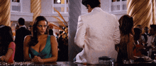 a woman in a blue dress sits at a bar with a man in a white suit behind her