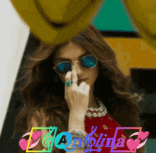a woman wearing sunglasses and a ring holds her finger to her nose in front of a colorful sign that says danolina