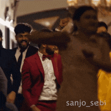 a man in a red suit and bow tie is dancing in a crowd with the name sanjo_sebi on the bottom