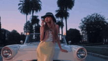 a woman in a hat is standing next to a white car with palm trees in the background