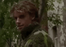 a man in a camouflage jacket is standing in the woods looking at the camera .