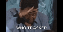 a man in a blue shirt is covering his face with his hand and the words `` who tf asked '' behind him .