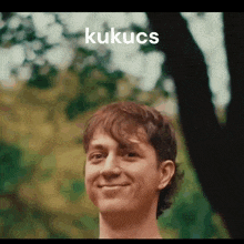 a young man is smiling in front of a tree and the word kukucs is on the bottom of his face .
