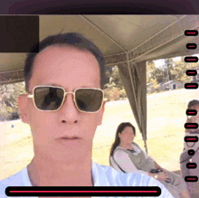a man wearing sunglasses takes a selfie with a woman sitting in the background