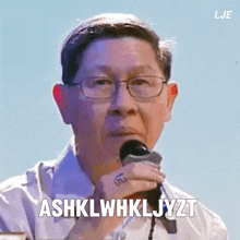 a man with glasses is holding a microphone and says ashklwhkljyzt