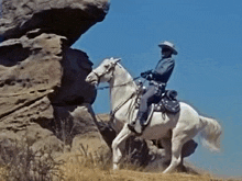 a man in a cowboy hat is riding a white horse on top of a rock .