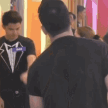 a man in a tuxedo is talking to another man in a black shirt in a crowded room .