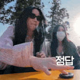 a woman wearing sunglasses and a face mask holds her hand out in front of another woman wearing a face mask