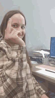 a woman in a plaid shirt is sitting at a desk with a computer in the background