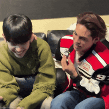 two men are sitting on a couch with one wearing a red jacket with the number 93 on it