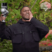 a man in a black jacket is holding a camera in front of a sign that says ultimate