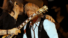 a man in a cowboy hat holds a guitar while another man looks on