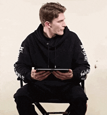 a man wearing a black hoodie with chinese writing on the sleeves is sitting on a chair holding a tablet