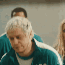a man with white hair and a green jacket is standing in a room with other people .