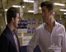 two men are standing next to each other and talking in a hallway .